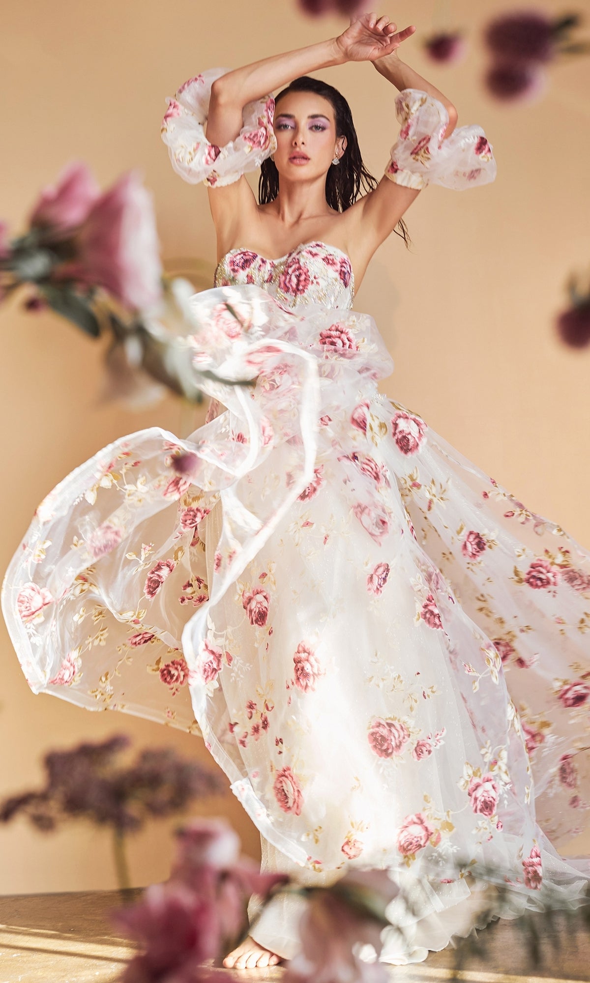 A woman poses barefoot in the Puff-Sleeve Strapless Floral-Print Ball Gown A1133, featuring sheer fabric and pink and white patterns, surrounded by blurred flowers against a beige background.